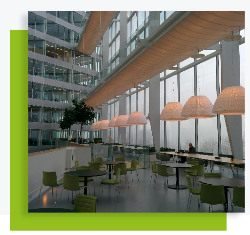 Salle lumineuse avec grandes baies vitrées, tables, chaises vertes et luminaires suspendus